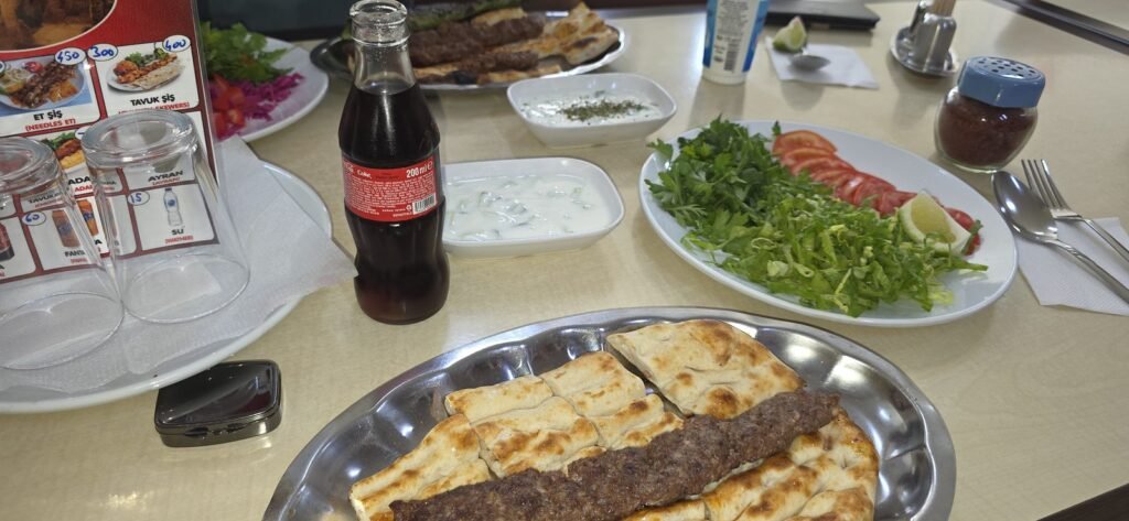 Voller Tisch bei Ali Usta in Derinkuyu mit Kebap, Cacık, Salat und Getränken, im Hintergrund die Preistafel